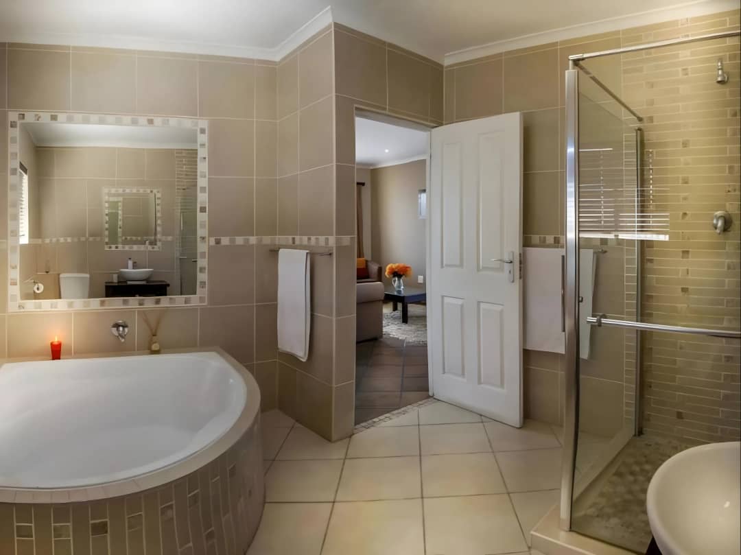 Tiled bathroom with corner tub, glass shower, mirror and open door to lounge.